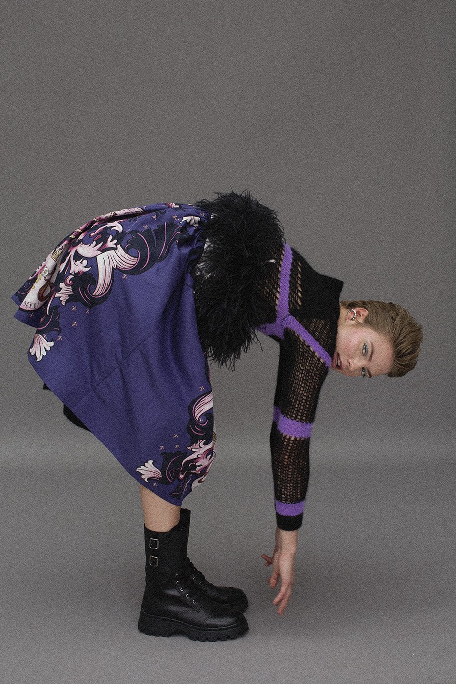 Woman in a black knit top and patterned skirt bends forward with black boots, set against a neutral grey backdrop in a studio.