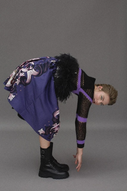 Woman in a black knit top and patterned skirt bends forward with black boots, set against a neutral grey backdrop in a studio.