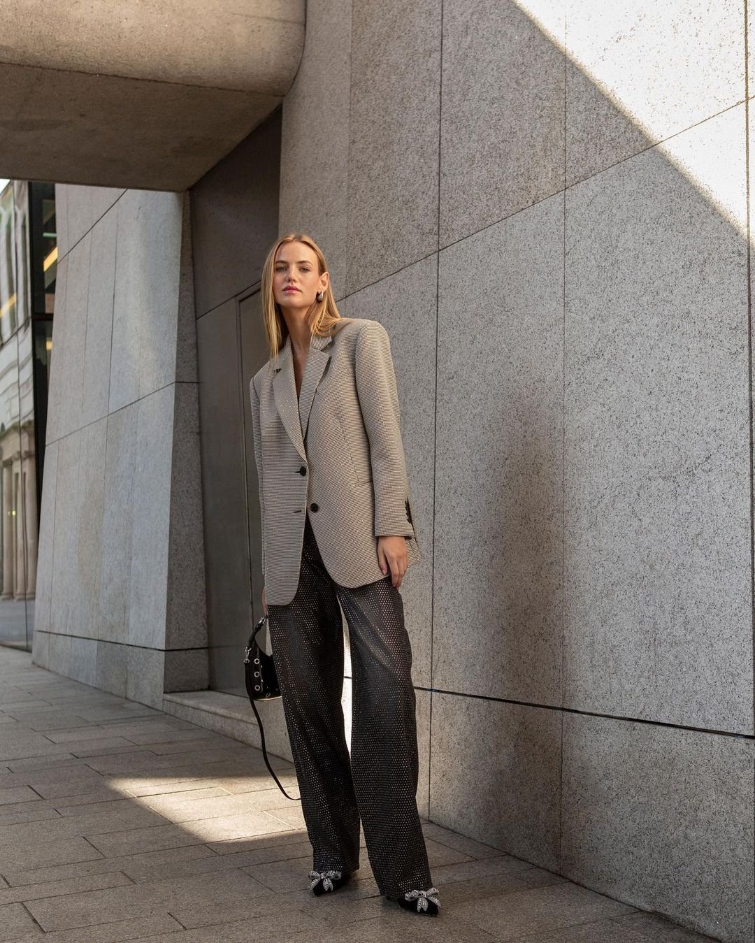 Stylish woman in an oversized blazer and sparkling pants stands in an urban setting with natural light highlighting her outfit.