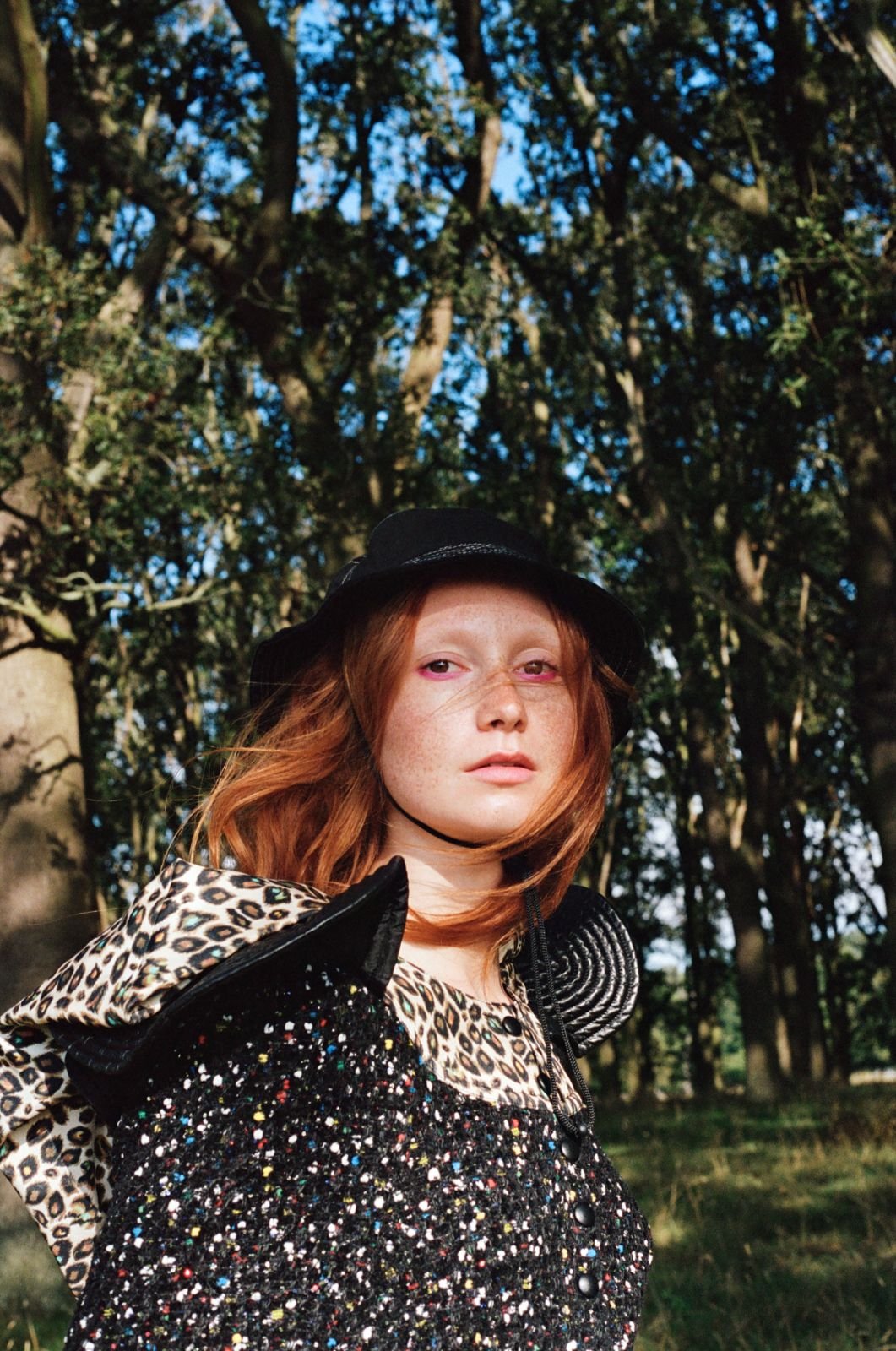 Portrait of a redhead woman wearing a hat and patterned top, standing outdoors against a backdrop of trees and natural light.