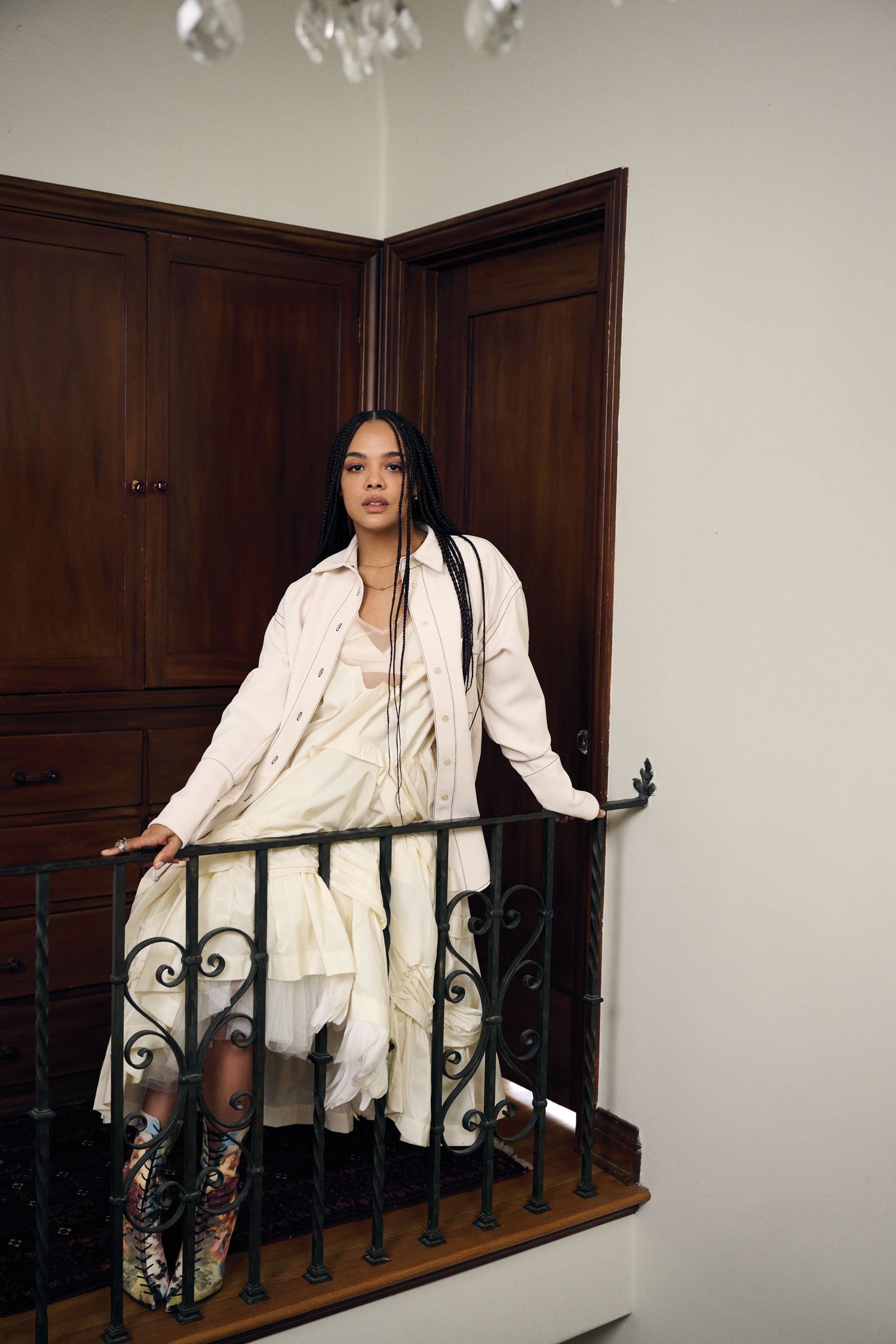 A woman with braids in a dress and jacket poses on a staircase with a wrought-iron railing inside of a home with brown wood paneling.