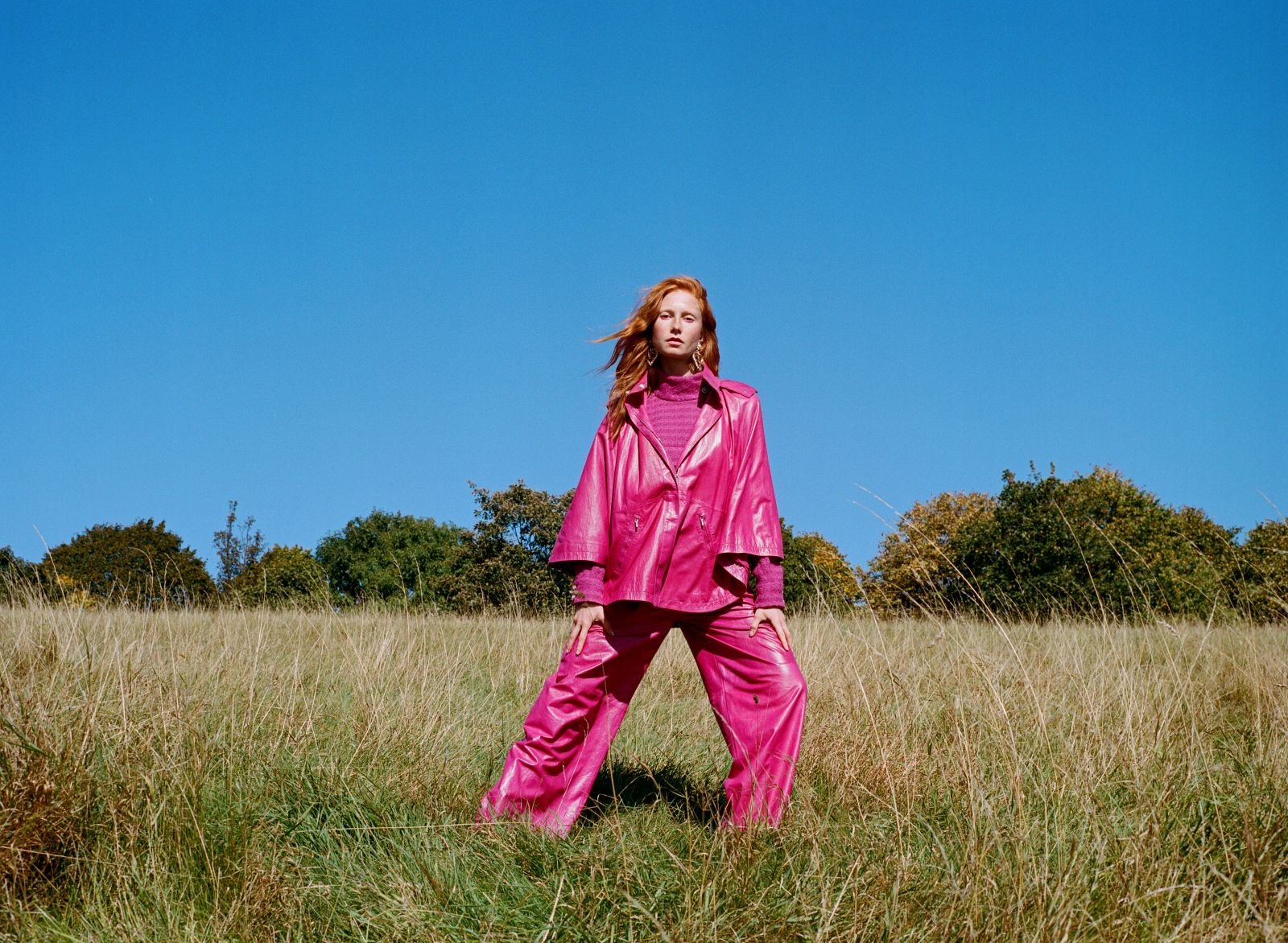 Full-length outdoor shot of red-haired woman in pink jacket and pants standing in a field under a bright blue sky, looking at camera.