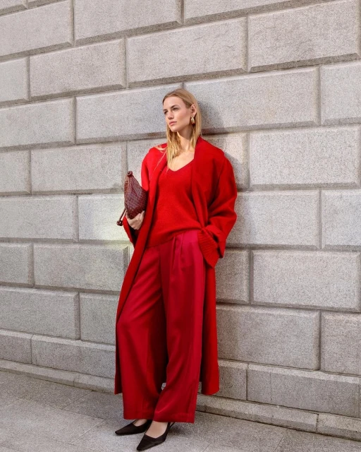 Stylish blonde woman poses in a red coat, red sweater, and red pants with a brown purse against a grey brick wall outdoors.