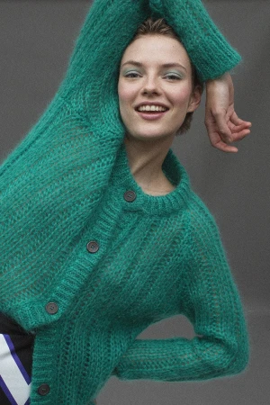 Smiling woman in a bright green mohair cardigan with arms raised against a plain grey backdrop.