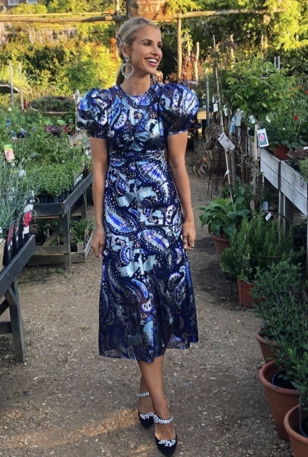 A smiling adult female stands outdoors in a stylish patterned midi dress and embellished heels, surrounded by potted plants.