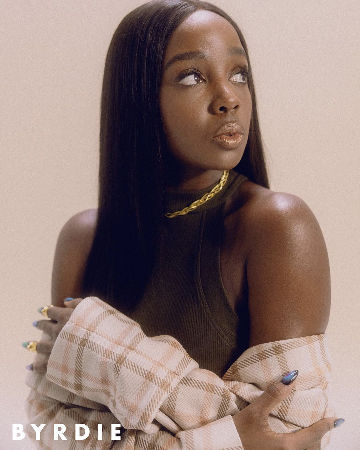 Portrait of a young woman with long black hair and gold necklace, wearing a tan plaid jacket, against a cream background.