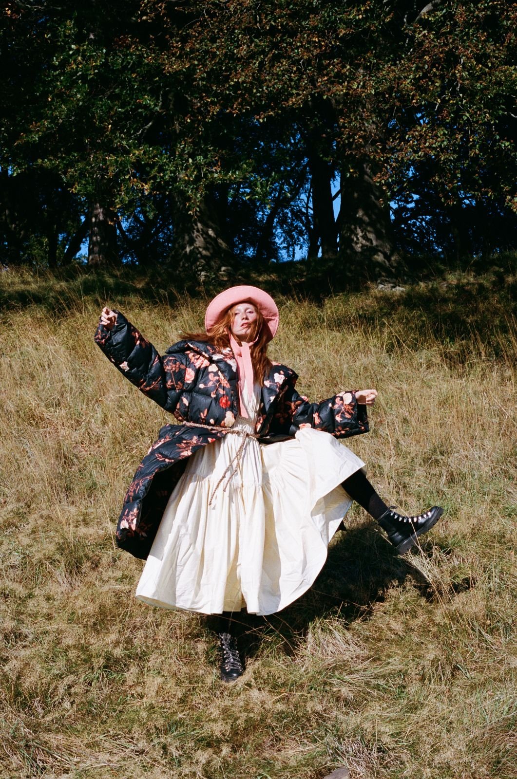 A woman in a floral coat, pink hat, and combat boots poses in a grassy field surrounded by trees on a sunny day in an outdoor fashion shoot.