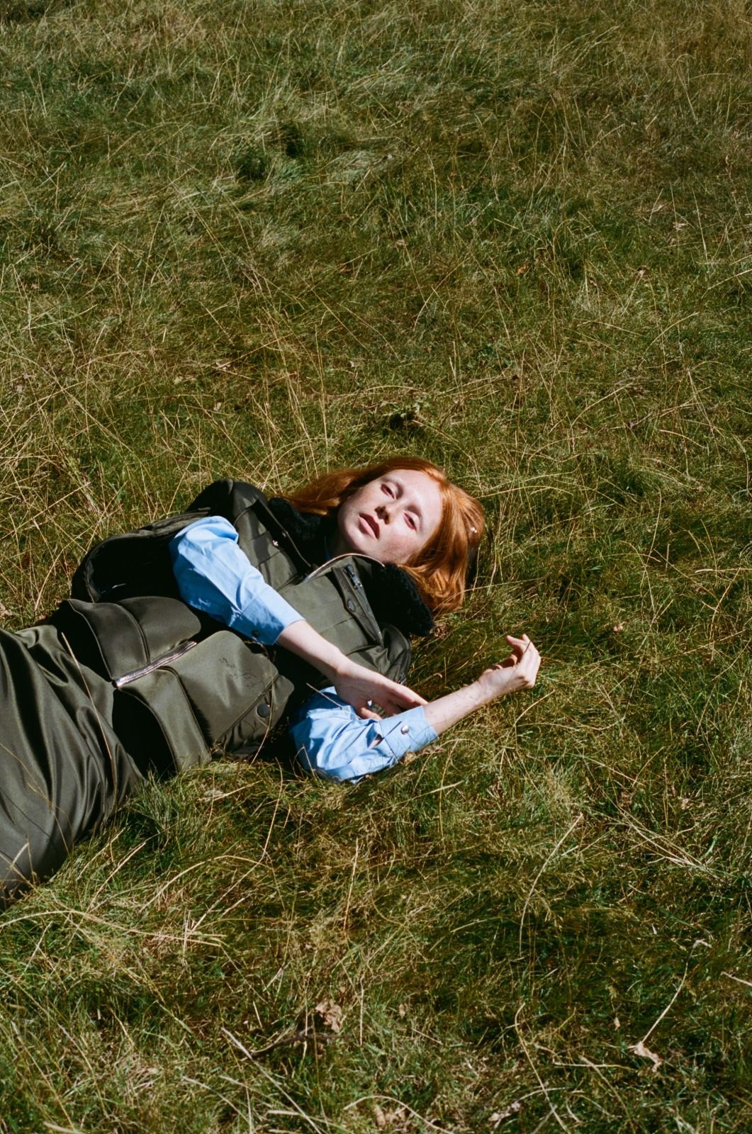 Red-haired woman in a vest and blouse lies on her back in dry grass in full sunlight, eyes open, her arm extending to the side.