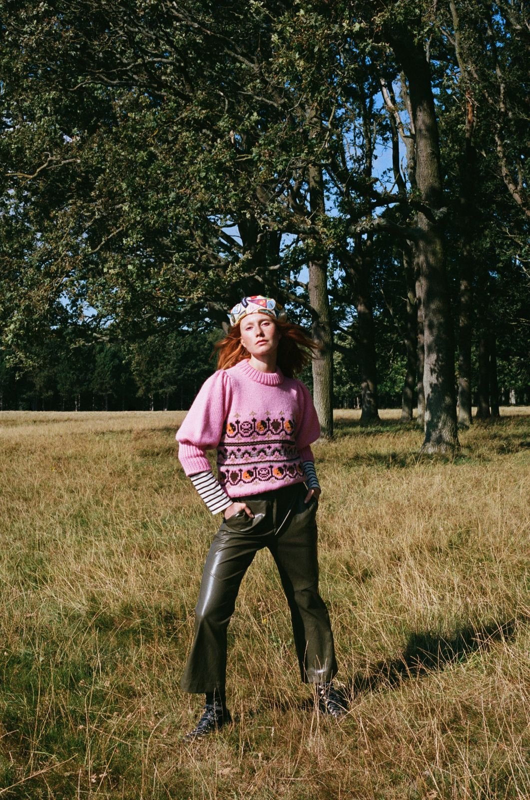 A woman in a pink sweater, leather pants, and floral headband stands in a grassy field with trees in the background on a sunny day.