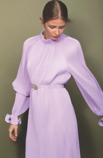 A woman wears a long-sleeved, light purple, pleated dress with a belt against a muted green backdrop in a fashion studio setting.