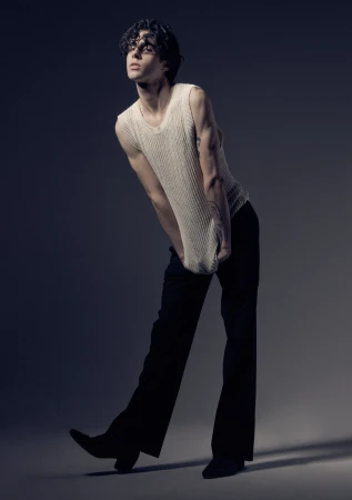 Young man with dark curly hair wearing a cream ribbed knit tank top and dark trousers posed against grey studio backdrop.