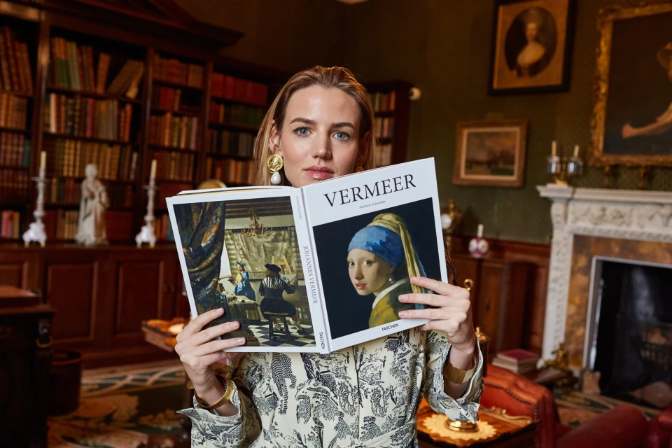 Adult woman holds a book featuring Johannes Vermeer's "Girl with a Pearl Earring" painting in an ornate room with a bookshelf.