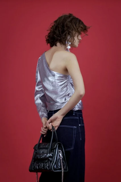 Fashionable woman in a silver asymmetrical top and dark jeans holds a structured black bag against a vivid red backdrop.