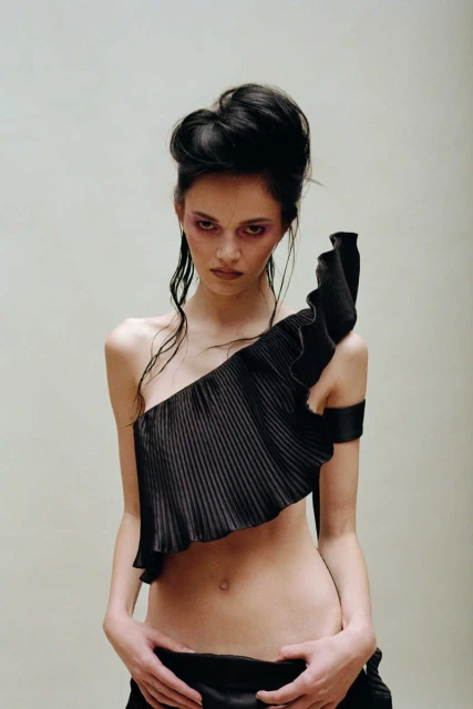 Fashionable woman in an avant-garde black top with flowing hair, posing in front of a neutral backdrop, showcasing an editorial aesthetic.