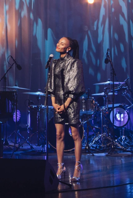 Full shot of a Black woman singing on stage in a sparkling dress, silver high heels, microphone in front of her, with a drum kit behind.