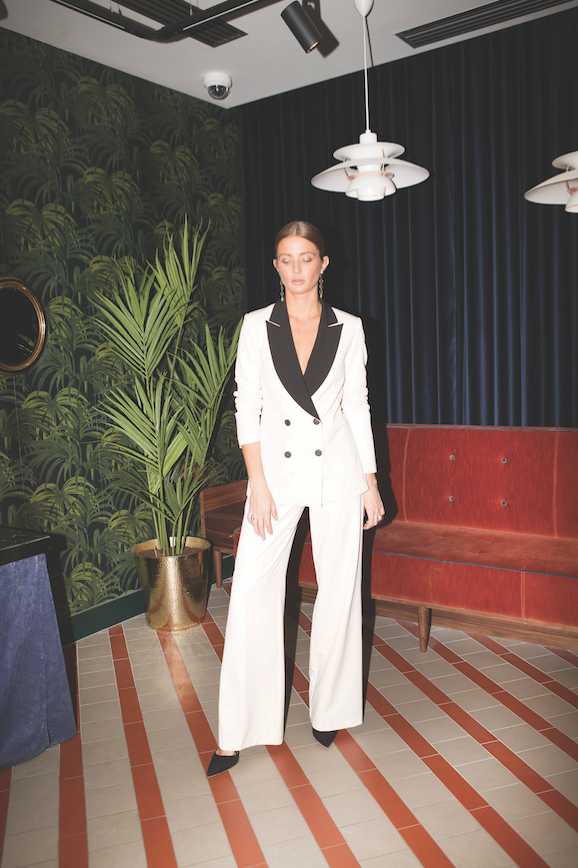 A woman in a white suit with black lapels strikes a pose indoors, standing on a tiled floor with a patterned wall and red couch in the background.