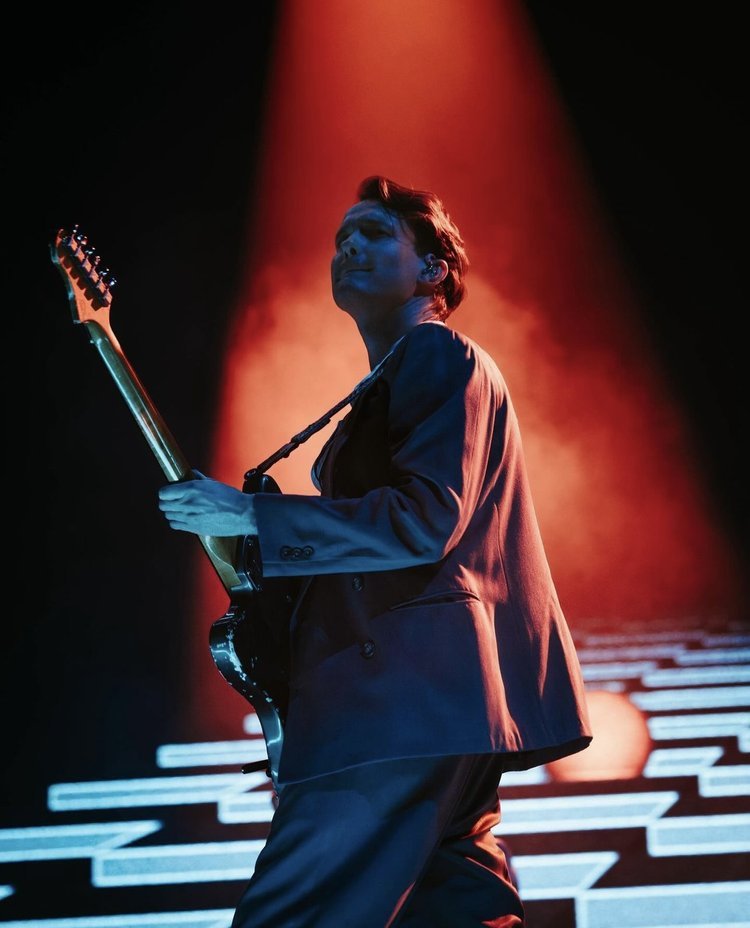 Man in a suit plays an electric guitar on stage under a spotlight with geometric set design; musician performing live at concert.
