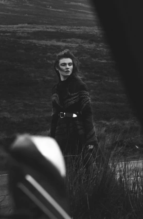 Monochrome fashion portrait of a model outdoors in a patterned coat standing near tall grass.