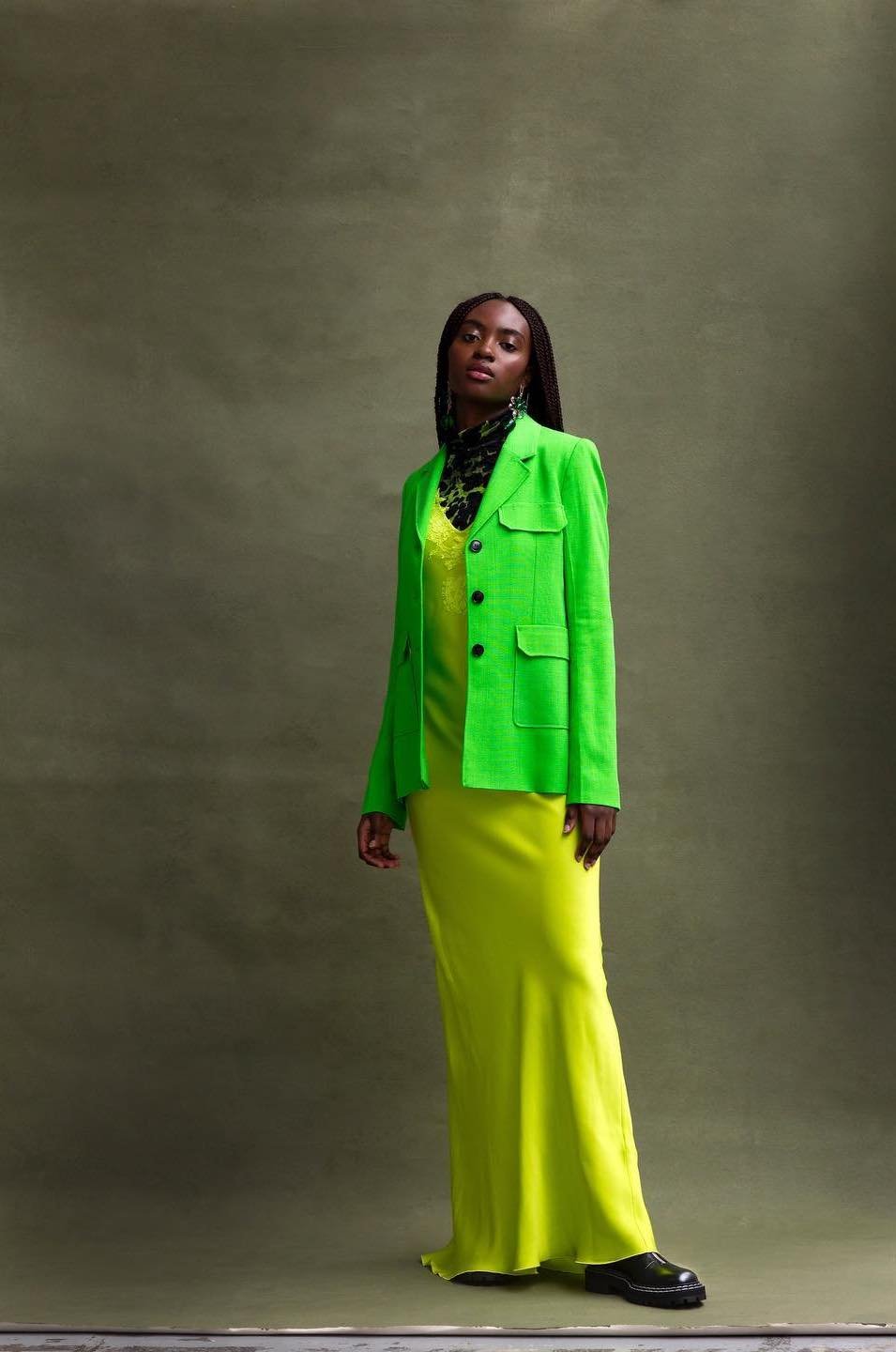 Full-length shot of a Black female model in a bright green blazer and yellow slip dress, posing against a mottled green backdrop.