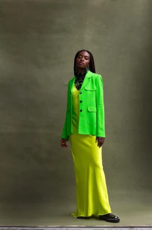 Full-length shot of a Black female model in a bright green blazer and yellow slip dress, posing against a mottled green backdrop.