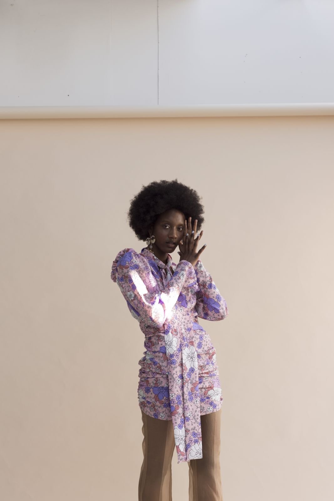 Black woman wearing a purple floral patterned long-sleeve top and brown trousers posing against a light neutral background.