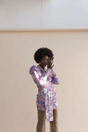 Black woman wearing a purple floral patterned long-sleeve top and brown trousers posing against a light neutral background.