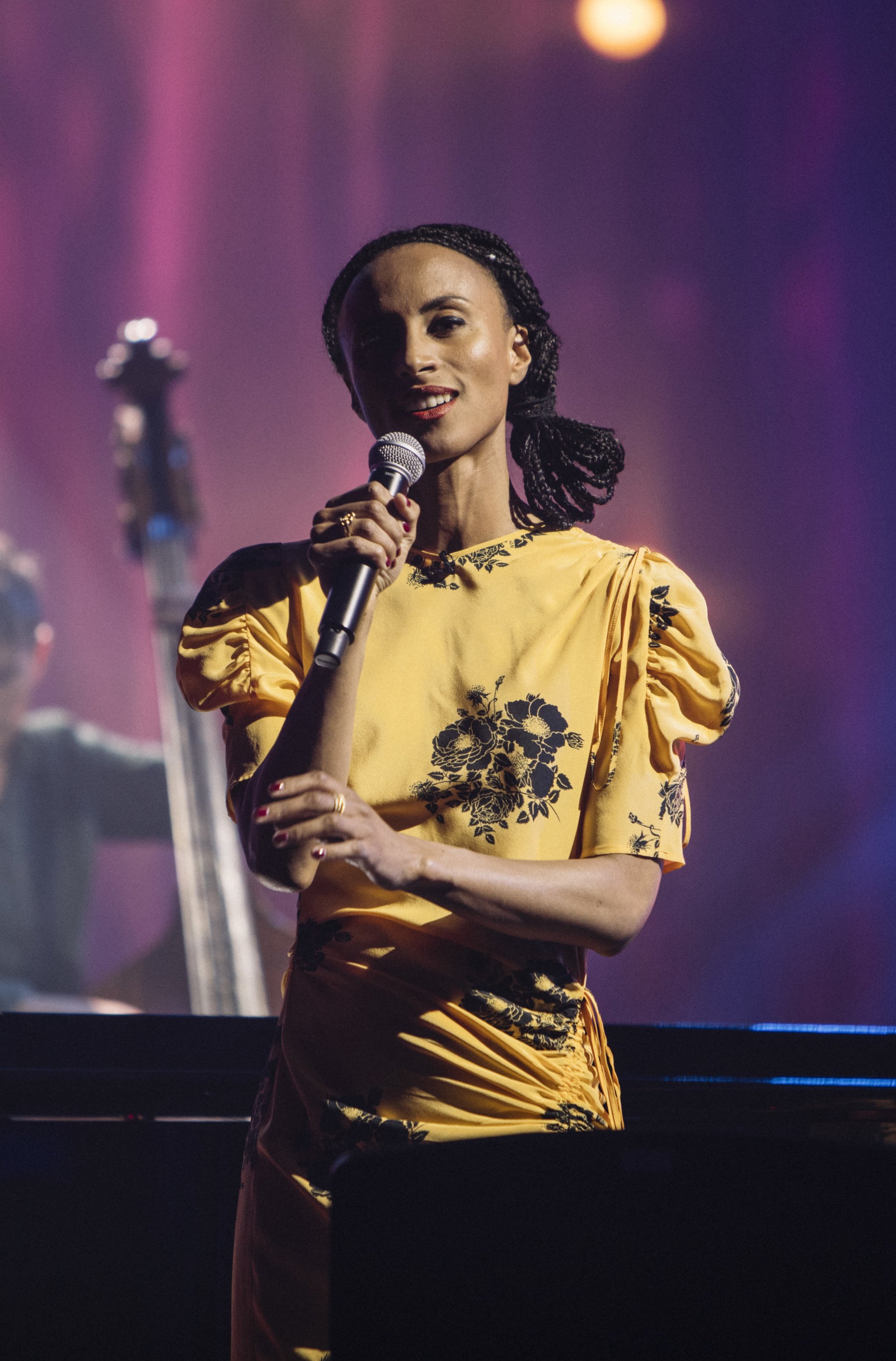An adult female singer in a yellow dress holds a microphone on a stage with musical instruments and pink and purple lighting.