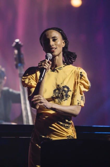 An adult female singer in a yellow dress holds a microphone on a stage with musical instruments and pink and purple lighting.