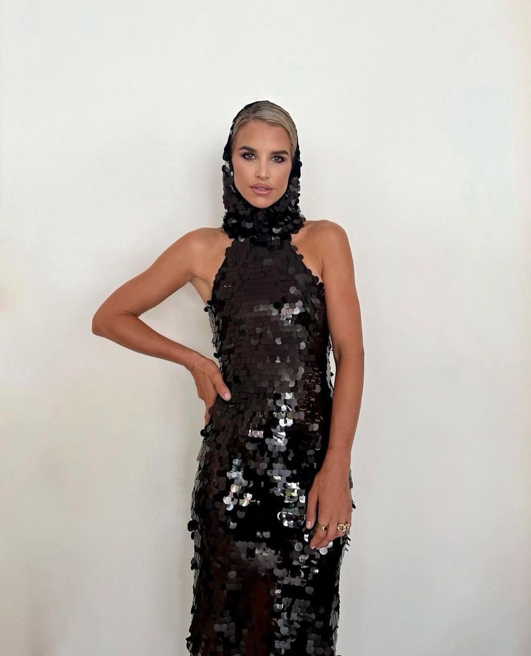 Woman in sequin dress poses against a white wall. Headpiece with sequins frames her face in a bright studio setting.