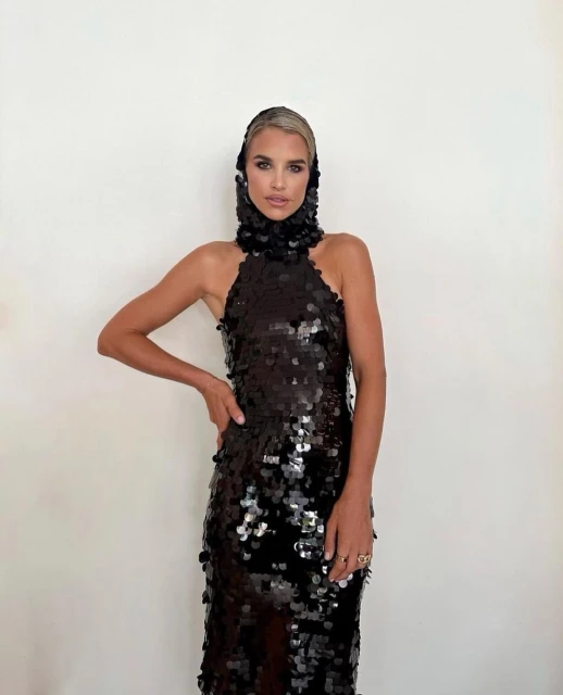 Woman in sequin dress poses against a white wall. Headpiece with sequins frames her face in a bright studio setting.