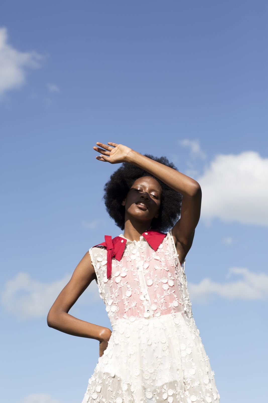 A dark-skinned woman poses outdoors, shielding her eyes from the sun. She wears a white dress with red accents against a blue sky backdrop.