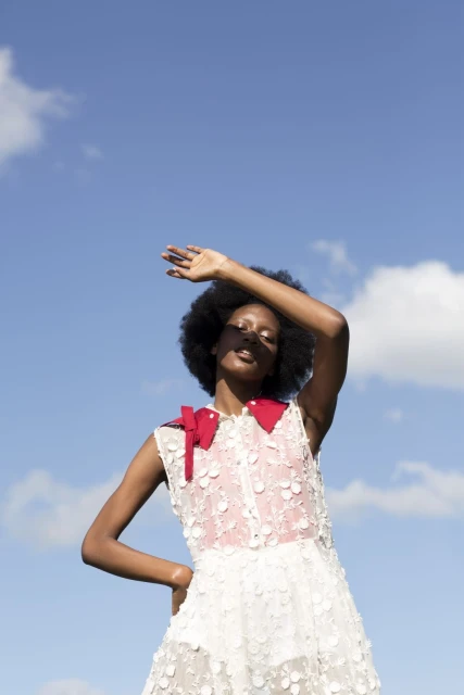 A dark-skinned woman poses outdoors, shielding her eyes from the sun. She wears a white dress with red accents against a blue sky backdrop.