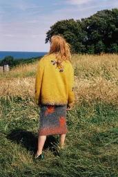 Woman with long blonde hair seen from behind wearing a yellow cardigan and grey patterned skirt standing in a coastal meadow