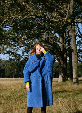 Red-haired model wearing a blue fluffy teddy coat standing outdoors in a grassy park beneath large trees