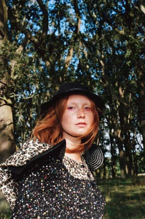 Red-haired model wearing a black bucket hat and textured tweed garment outdoors among dense trees.