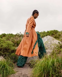 Model in a long caramel coat and patterned green trousers walking among rocky terrain and green foliage.