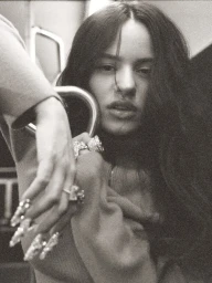 Monochrome portrait of a woman with long dark hair, wearing ornate rings and long embellished fingernails, holding a metal grab handle.