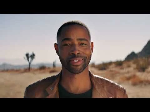 Close-up shot of an African-American adult male with a beard, smiling in a desert landscape with mountains and a blue sky.