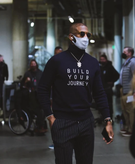 Adult man with sunglasses and face mask wears a navy turtleneck sweater with white text, striped pants, and walks in an indoor setting.