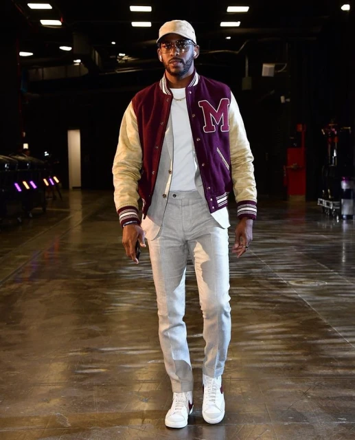 Stylish adult male in letterman jacket, grey suit, and white sneakers, walking indoors on a glossy floor with dark background lighting.