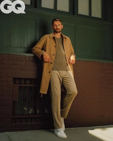Man wearing a tan trench coat, brown cable-knit sweater, and trousers leaning against a dark green and brick wall.