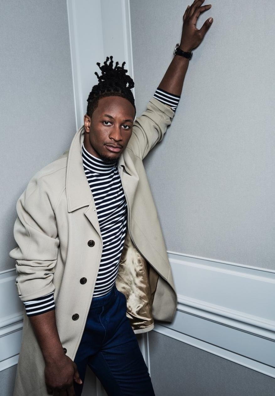 Man with dreadlocks poses in a beige trench coat and striped turtleneck indoors against a paneled wall, arm raised, looking at the camera.