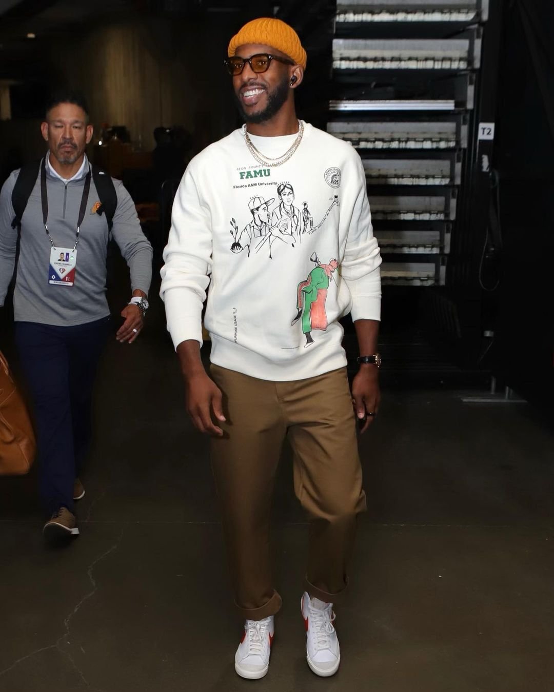 Adult male in an orange beanie and sunglasses wearing a FAMU sweatshirt and khaki pants walks toward the camera on an indoor walkway.