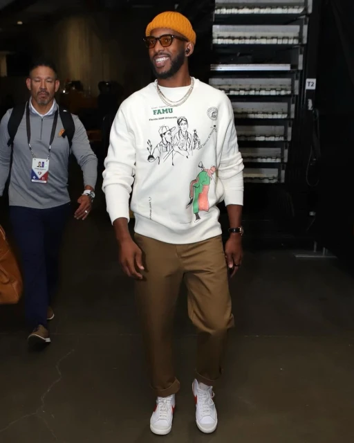 Adult male in an orange beanie and sunglasses wearing a FAMU sweatshirt and khaki pants walks toward the camera on an indoor walkway.