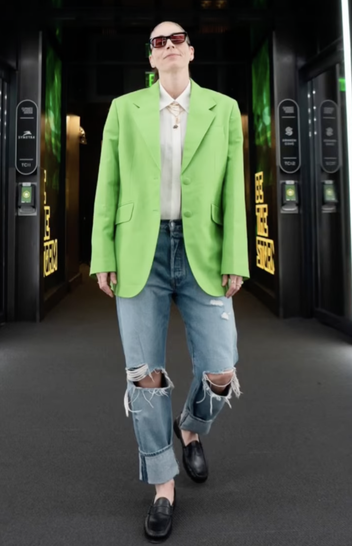 A woman in sunglasses, a neon green blazer, and ripped blue jeans stands in a corridor with decorative dark-green paneled walls.