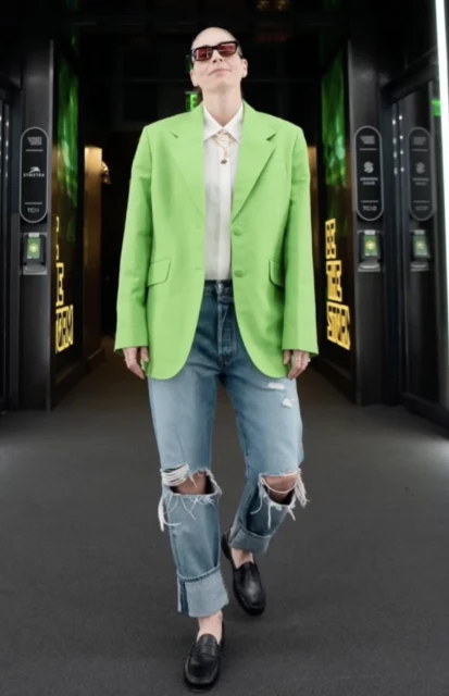A woman in sunglasses, a neon green blazer, and ripped blue jeans stands in a corridor with decorative dark-green paneled walls.