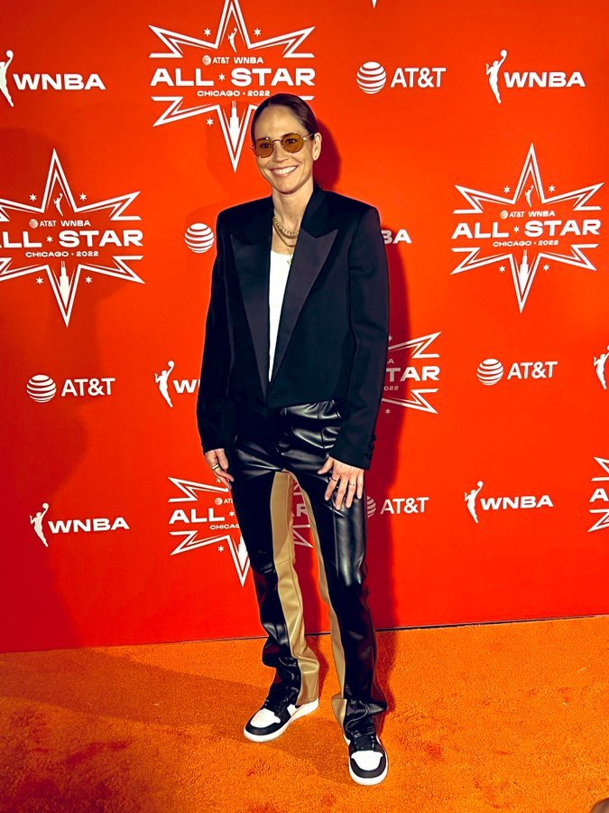 A woman in a blazer and leather pants poses on an orange carpet at the WNBA All-Star event in Chicago. The background features sponsor logos.