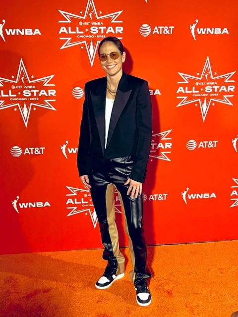 Adult female in a black blazer, white top, and black pants posing at the WNBA All-Star event on an orange branded backdrop.