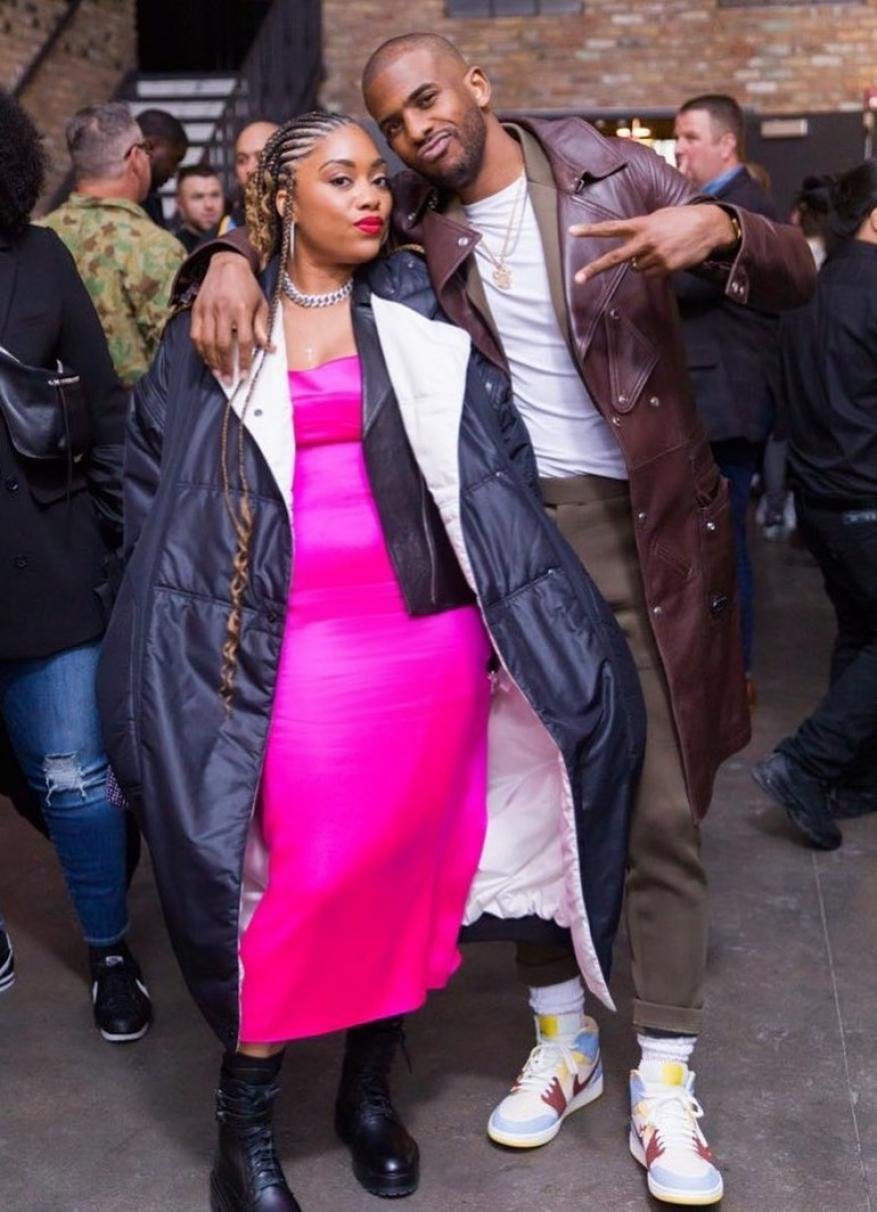 Stylish couple posing indoors: Woman in bright pink dress and black coat, man in brown leather jacket, standing close together in a group.