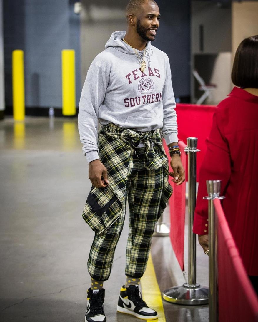 Man in casual clothing, including a hoodie and plaid pants, stands in an indoor setting with yellow pillars.